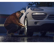 Someone is changing a tire on a white car in a dimly lit area, wearing a brown jacket and using the Armytek Elf C1 USB-C White headlamp, which provides 1000 lumens of light to ensure ample illumination for the task.