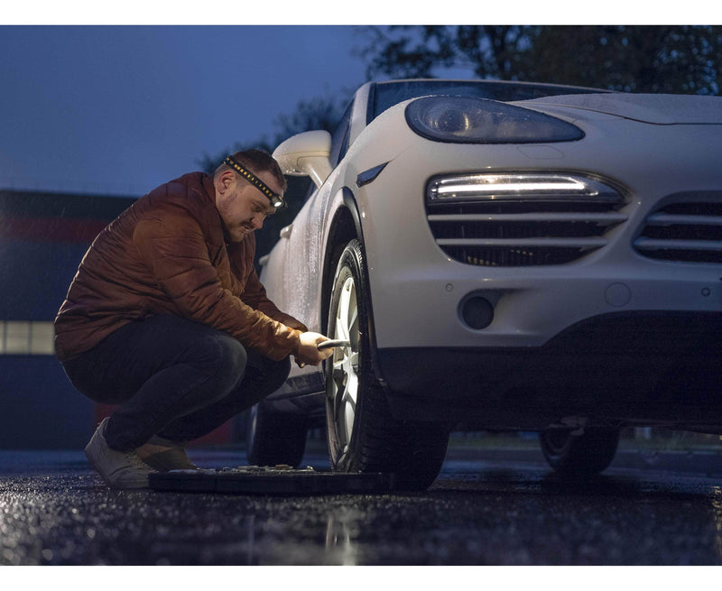 Someone is changing a tire on a white car in a dimly lit area, wearing a brown jacket and using the Armytek Elf C1 USB-C White headlamp, which provides 1000 lumens of light to ensure ample illumination for the task.