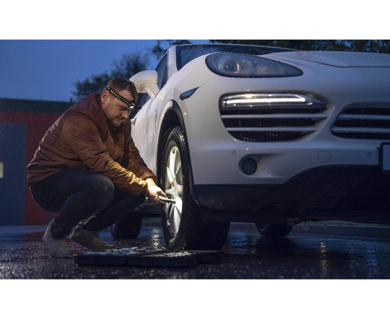 Using the Armytek Elf C2 USB-C Warm headlamp with 1023 lumens, an individual changes a tire on a white SUV at dusk as its light cuts through the rain.