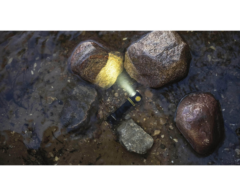 The Armytek Elf C2 USB-C Warm flashlight, featuring a robust design and USB-C charging capability, is submerged in shallow water among rocks, emitting a steady beam of light.