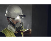 In a dimly lit environment, a person wearing an Armytek hard hat with goggles and a respirator points at a surface. The Armytek Elf C2 USB-C Warm headlamp attached to the helmet shines brightly, while the convenient USB-C connection ensures it stays charged and ready for any task.