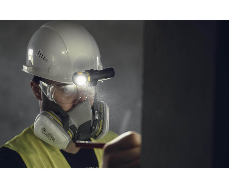 In a dimly lit environment, a person wearing an Armytek hard hat with goggles and a respirator points at a surface. The Armytek Elf C2 USB-C Warm headlamp attached to the helmet shines brightly, while the convenient USB-C connection ensures it stays charged and ready for any task.