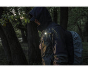 A person wearing a dark jacket and hood walks through a forest with a backpack, illuminated by the Armytek Elf C2 USB-C Warm flashlight that shines brightly at 1023 lumens.
