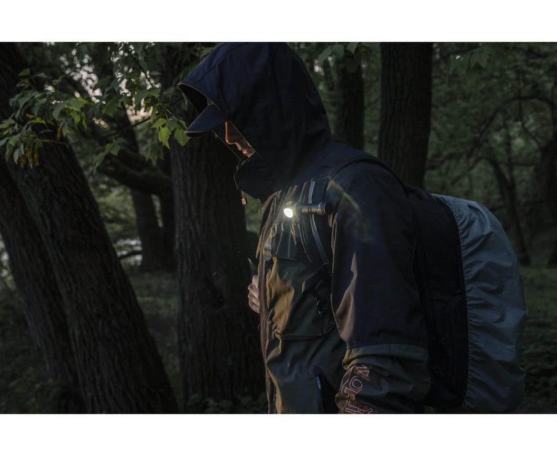 A person wearing a dark jacket and hood walks through a forest with a backpack, illuminated by the Armytek Elf C2 USB-C Warm flashlight that shines brightly at 1023 lumens.
