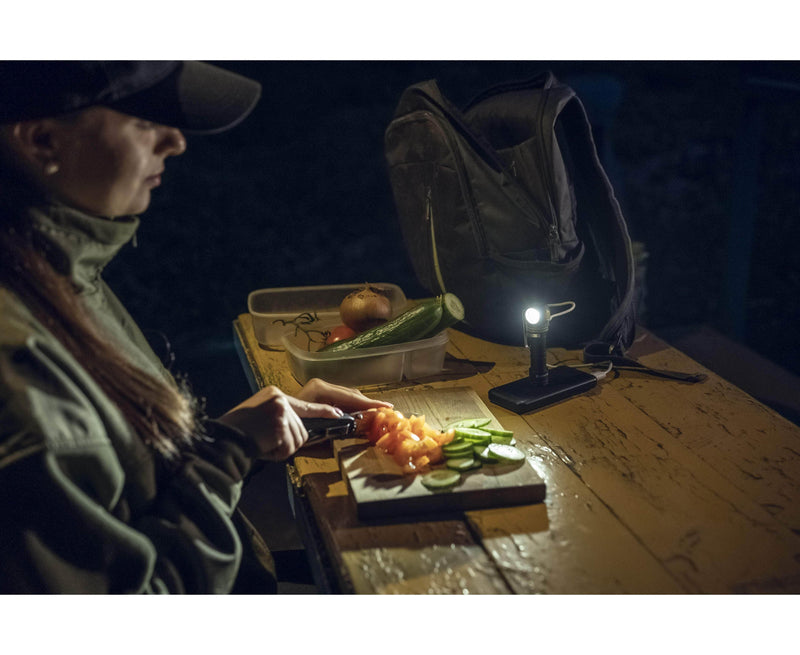 A person is cutting vegetables by the light of their Armytek Elf C2 USB-C Warm, casting a warm glow over the wooden table. Their backpack and food container sit ready, alongside a USB-C charger, ensuring everything stays powered through the night.