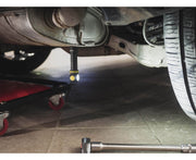 An Armytek Elf C2 USB-C Warm flashlight illuminates the underside of a vehicle in a mechanic's workshop, with a wheeled creeper and a wrench nearby. The scene suggests efficiency and modernity, enhanced by tools featuring USB-C ports for quick charging.