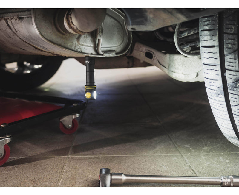 An Armytek Elf C2 USB-C Warm flashlight illuminates the underside of a vehicle in a mechanic's workshop, with a wheeled creeper and a wrench nearby. The scene suggests efficiency and modernity, enhanced by tools featuring USB-C ports for quick charging.