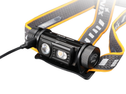 The Fenix HM60R is an outdoor headlamp in black and orange, equipped with a strap and two LED lights capable of delivering up to 1300 lumens. An additional cable connects to the side, enhancing its functionality.