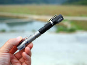 A hand holds the Manker E02 III - Titanium, a compact flashlight with a pocket clip that emits 560 lumens—ideal for everyday carry—displayed outdoors near a blurred body of water and grassy landscape.