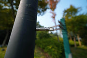 Close-up of a metallic bolt attached to a dark cylindrical pole outdoors, echoing the sleek design of the Manker E02 III - Titanium flashlight, with softly blurred green trees and foliage in the background.