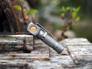 The Manker E02 III - Titanium, a compact flashlight with a textured grip, sits on weathered wooden planks outdoors amidst blurred greenery—ideal for everyday carry.