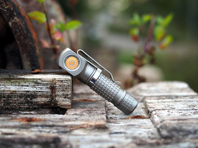 The Manker E02 III - Titanium, a compact flashlight with a textured grip, sits on weathered wooden planks outdoors amidst blurred greenery—ideal for everyday carry.