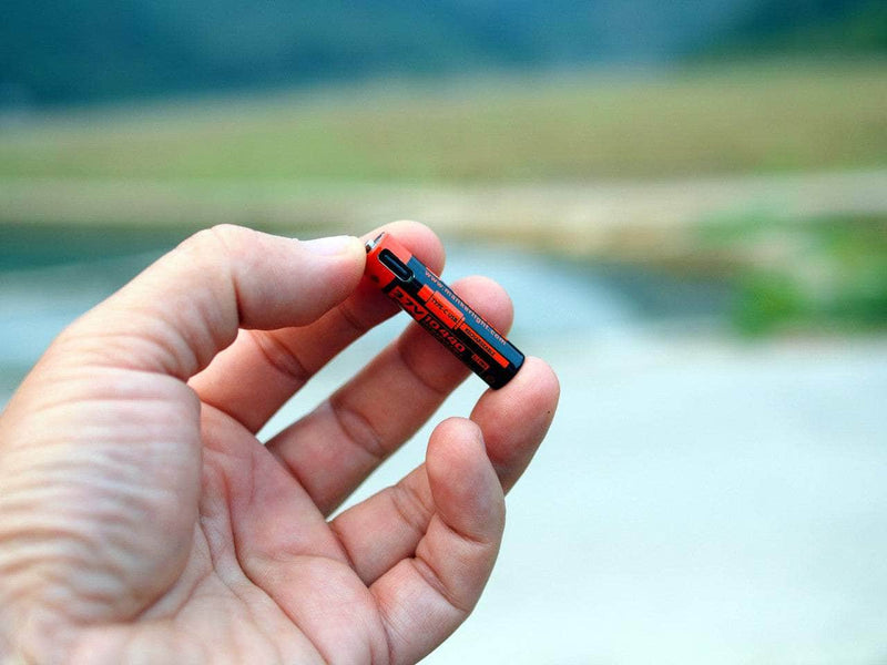 A hand holds a small black and red AA battery outdoors with a blurred natural background—ideal for powering your Everyday Carry, such as the Manker E02 III - Titanium flashlight.