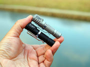 A hand holds two compact, textured Manker E02 III - Titanium flashlights—one black and one silver—over a blurred outdoor background near water, ideal for everyday carry with up to 560 lumens of brightness.