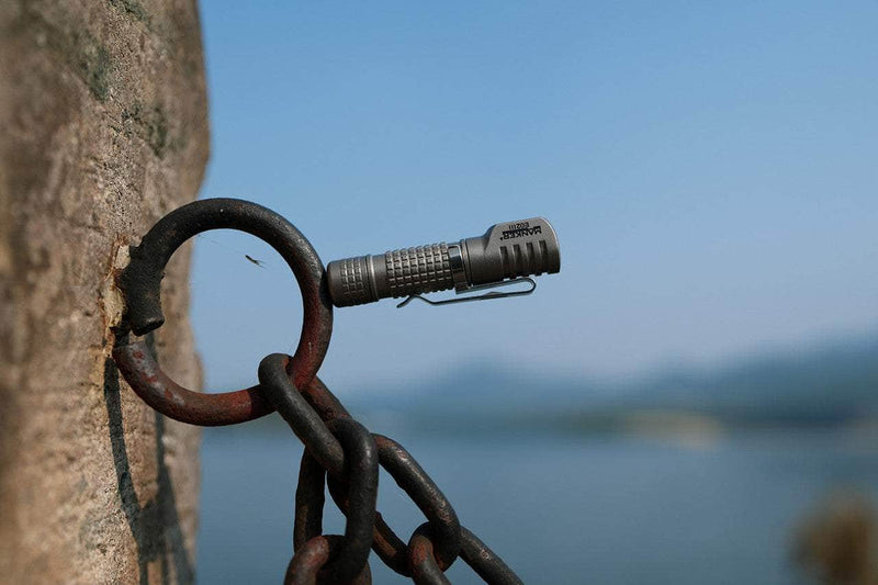 The Manker E02 III - Titanium flashlight, clipped to a rusty metal ring on a chain, shines with a backdrop of blurred water and mountains—an ideal companion for everyday carry.