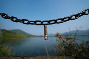 The Manker E02 III - Titanium flashlight hangs from a metal chain by the lake, with hills and greenery behind it under a clear blue sky—an ideal everyday carry companion.