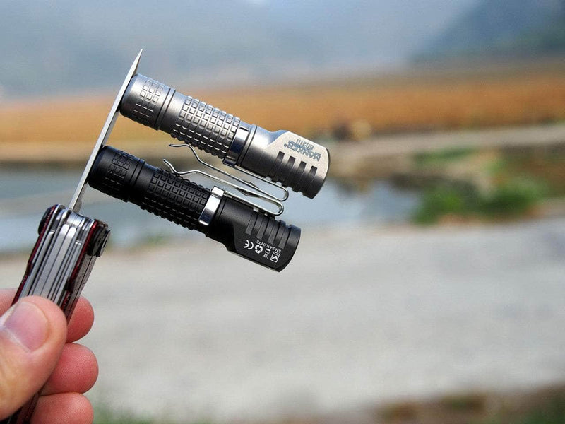A hand holds an everyday carry multitool with two compact flashlights attached, including the Manker E02 III - Titanium in silver and a black one. The 560 lumen brightness stands out against a blurred outdoor landscape backdrop.