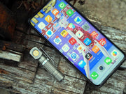 A smartphone with numerous app icons sits on a wooden surface next to the Manker E02 III - Titanium flashlight, offering 560 lumens—an essential everyday carry companion.