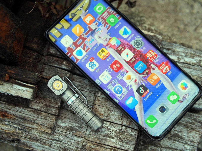 A smartphone with numerous app icons sits on a wooden surface next to the Manker E02 III - Titanium flashlight, offering 560 lumens—an essential everyday carry companion.