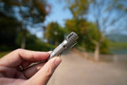 A hand holds the Manker E02 III - Titanium Flashlight outdoors on a paved path with trees and a blurred background, making it the ideal Everyday Carry for any adventure.