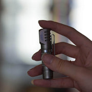 A hand holds the Manker E02 III - Titanium, a sleek flashlight with a textured grip and pocket clip—perfect for everyday carry—against a blurred background, ready to shine with 560 lumens of brightness.