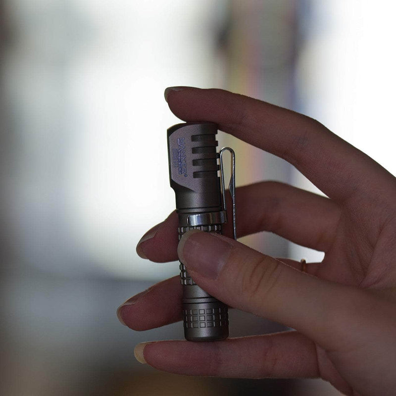 A hand holds the Manker E02 III - Titanium, a sleek flashlight with a textured grip and pocket clip—perfect for everyday carry—against a blurred background, ready to shine with 560 lumens of brightness.