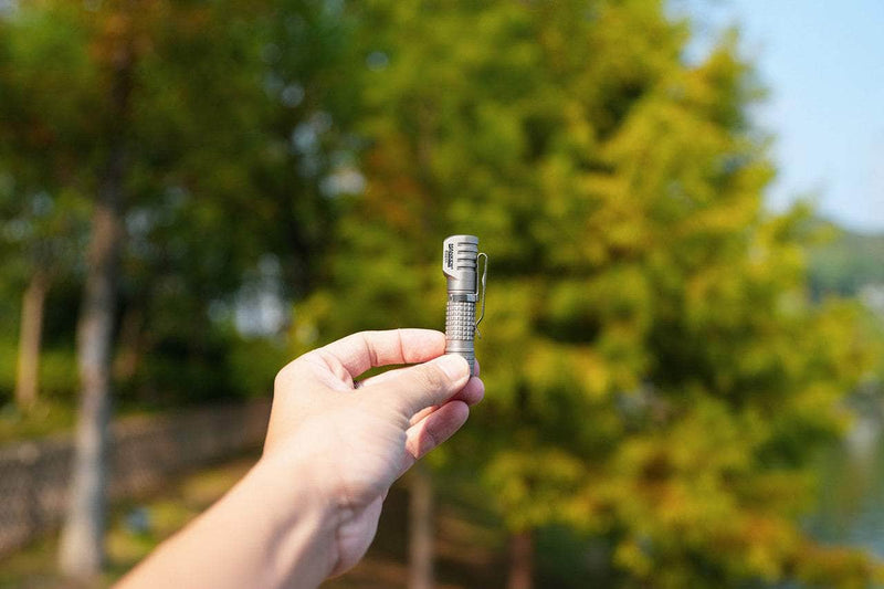 A hand holds the Manker E02 III - Titanium flashlight outdoors, with blurred green trees in the background—an ideal compact everyday carry tool.