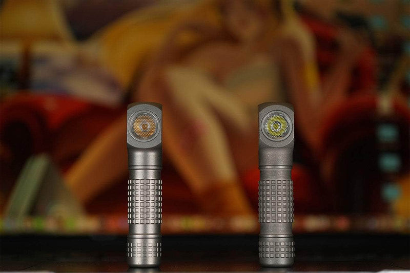 Two Manker E02 III - Titanium Everyday Carry Flashlights stand upright on a flat surface, providing 560 lumens of illumination, with a blurred painting of a person in the background.