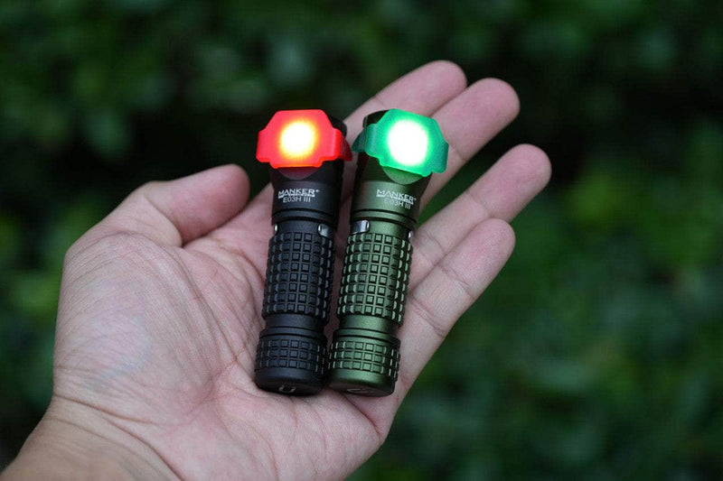 A hand holding two Manker E03H III everyday carry flashlights, one emitting red light and the other green, each featuring a textured grip design; trees form the background.