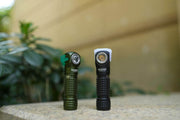 Two compact, angled Manker E03H III flashlights stand on a stone surface outdoors, surrounded by greenery and a blurred background. One 1200-lumens flashlight is green with black accents, and the other everyday carry flashlight is black with white accents.