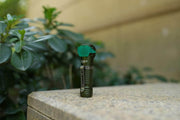 The Manker E03H III, in vibrant green, stands upright on a stone surface with green leaves and a wall in the background, making it perfect for everyday carry.