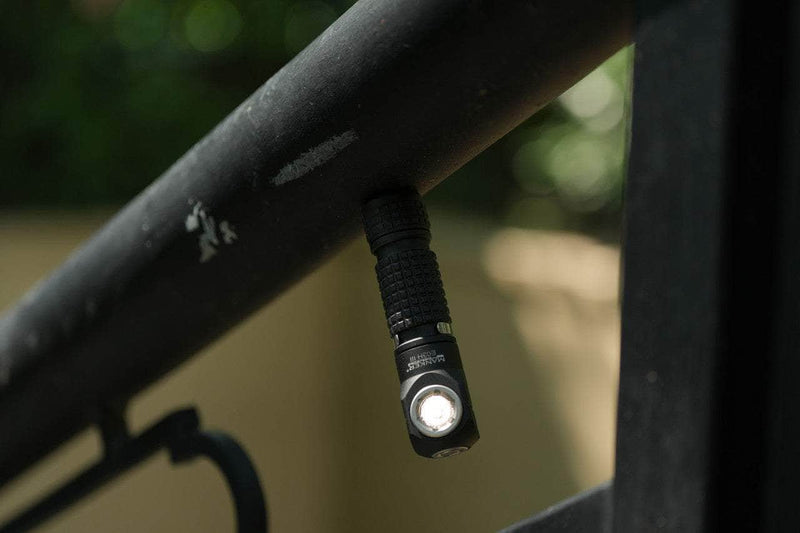 Close-up image of a Manker E03H III, a 1200 lumens everyday carry flashlight, hanging from a metal bar outdoors with a blurred background.