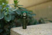 A Manker E03H III, a small green flashlight perfect for everyday carry, is placed on a stone surface outdoors, with green foliage in the blurred background.