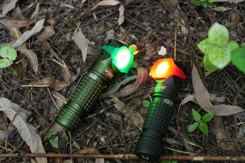 Two Manker E03H III everyday carry flashlights with red and green glowing ends, each boasting 1200 lumens, lie on a forest floor covered with leaves and small plants.