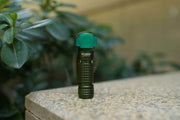 A small green and black device, identified as a "Manker E03H III," is placed on a stone surface with greenery in the background. Known for being one of the brightest everyday carry options, this versatile tool is perfect for outdoor adventures.