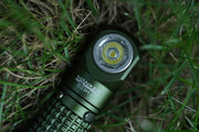 A Manker E03H III flashlight, boasting 1200 lumens, lies on the grass, showcasing its textured handle and circular LED light—an impressive everyday carry flashlight in a striking green color.