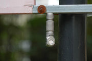 A compact titanium Manker E03H III is magnetically attached upside down to a metal bar next to a rusted bolt, with a soft-focus outdoor background.