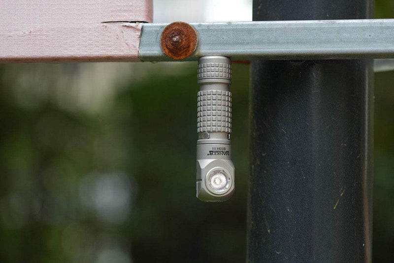 A compact titanium Manker E03H III is magnetically attached upside down to a metal bar next to a rusted bolt, with a soft-focus outdoor background.