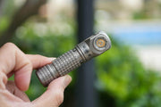 A hand holds the compact Manker E03H III - Titanium flashlight with an angled head outdoors, set against blurred greenery and sky; this light delivers a powerful 1200 lumens.