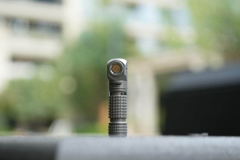A compact, metallic Manker E03H III - Titanium flashlight stands upright outdoors on a flat surface, with blurred greenery and buildings behind it—providing up to 1200 lumens of brightness.
