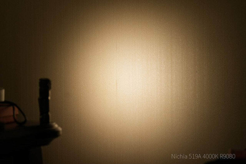 A Manker E03H III - Titanium flashlight casts a warm, circular 1200-lumen beam onto a beige wall in a dim room, with the flashlight partially visible on the left.