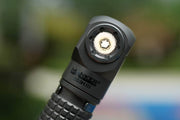 Close-up of a Manker E03H III - Titanium flashlight showing its LED emitter and textured grip, set against a blurred background. This compact light outputs a powerful 1200 lumens for impressive illumination.