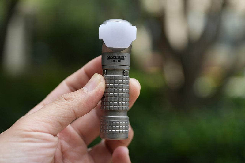 A hand holds the Manker E03H III - Titanium flashlight with a textured grip and white diffuser cap, outdoors against blurred greenery. This compact torch delivers up to 1200 lumens for powerful illumination.