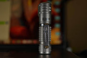 A compact Manker E03H III - Titanium flashlight stands upright on a dark surface with a blurred background, ready to deliver up to 1200 lumens of brightness.