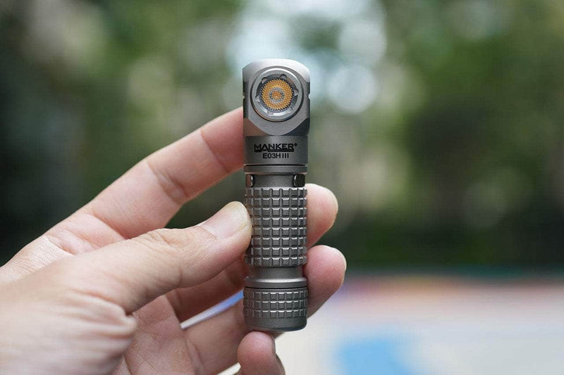 A hand holds the compact Manker E03H III - Titanium flashlight outdoors against a blurred green background; this powerful flashlight delivers up to 1200 lumens.