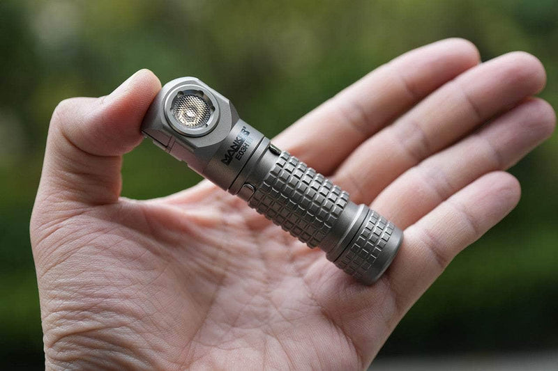 A person holds a compact Manker E03H III - Titanium flashlight in their hand, set against a blurred outdoor background.