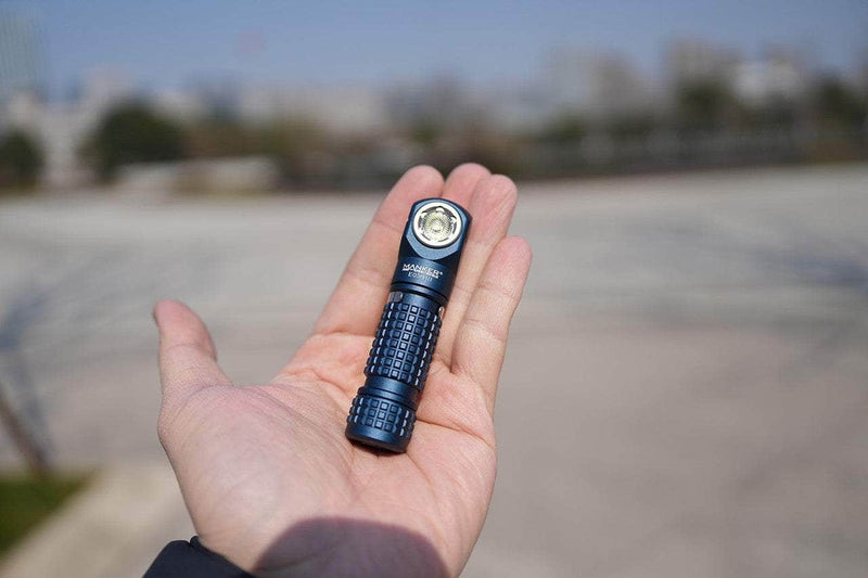 A person holds the compact Manker E03H III blue flashlight in their open hand outdoors, with a blurred background of trees and buildings, emphasizing its powerful lumens output and portable design.
