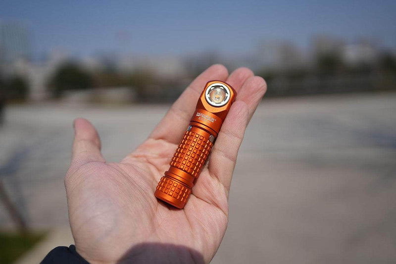 A person holds the orange Manker E03H III, a compact 1200-lumen right-angle flashlight, outdoors with buildings and trees blurred in the background—perfect for everyday carry.