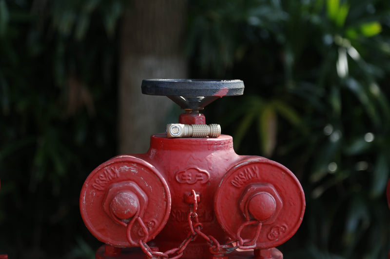 A red fire hydrant with two hose connections and a black valve handle, set outdoors against green foliage, is vividly illuminated by the Manker E03H III AA headlamp (1200 lumens), providing enhanced visibility.