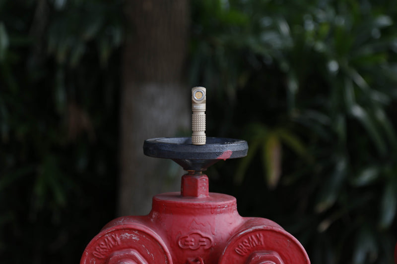 The compact beige Manker E03H III flashlight, boasting 1200 lumens, stands upright on its black metal cap atop a red fire hydrant, with green foliage and a tree trunk softly blurred in the background.